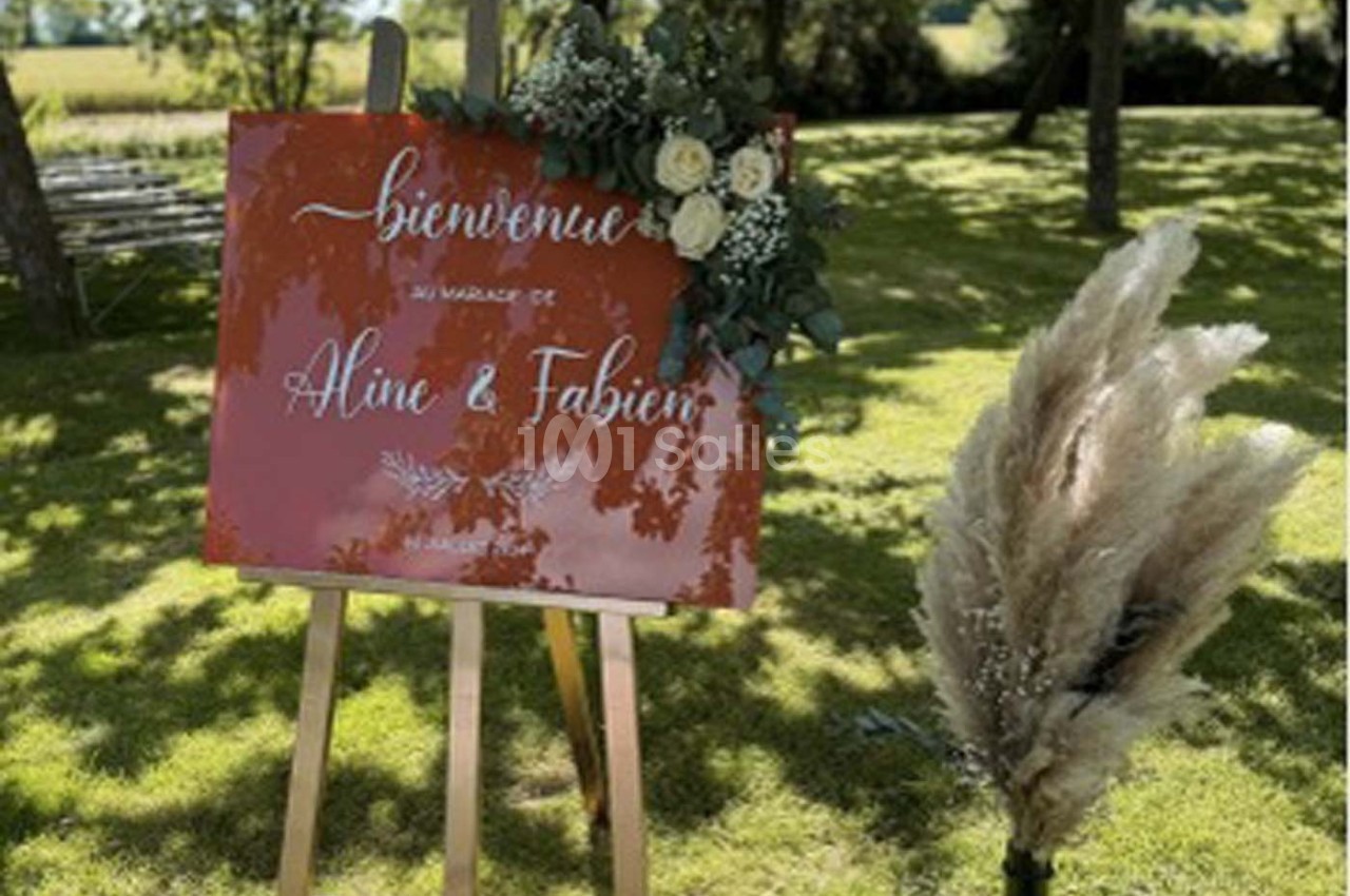 Panneau de bienvenue rouge sur un chevalet, décoré de fleurs blanches et de pampas, posé sur une pelouse ensoleillée.