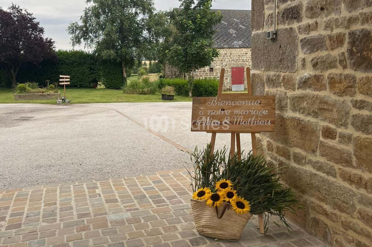 Panneau en bois indiquant ’Bienvenue à notre mariage Alice & Mathieu’ avec un panier de tournesols devant un bâtiment en…