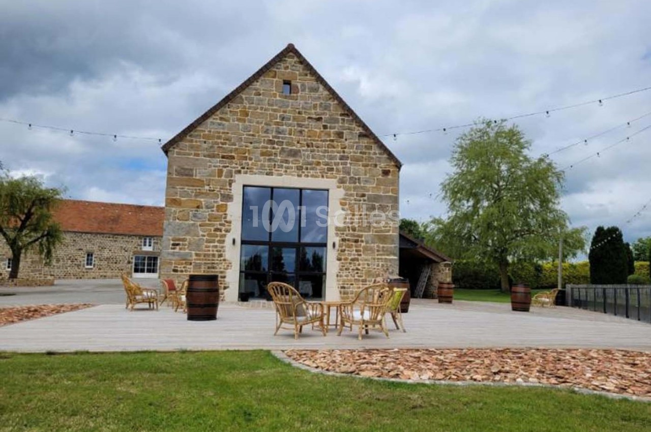 Bâtiment en pierre avec une grande baie vitrée, terrasse en bois aménagée de chaises et tables, entouré de verdure.