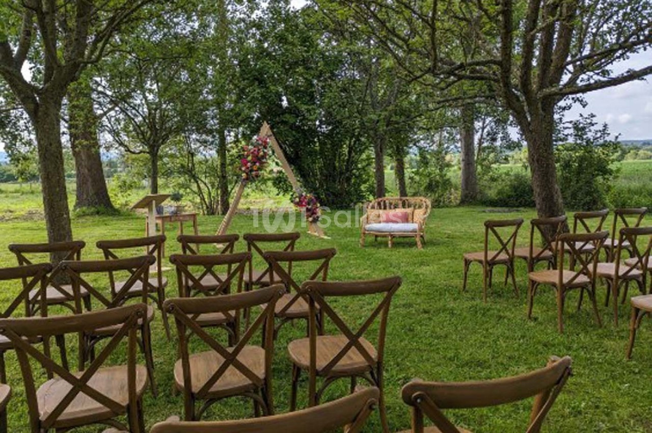Chaises en bois disposées en extérieur face à une arche décorée de fleurs, entourées d'arbres et de verdure.