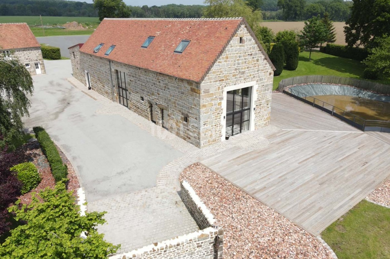 Bâtiment en pierre rénové avec toit en tuiles rouges, entouré d'une terrasse en bois et d'un paysage rural verdoyant.