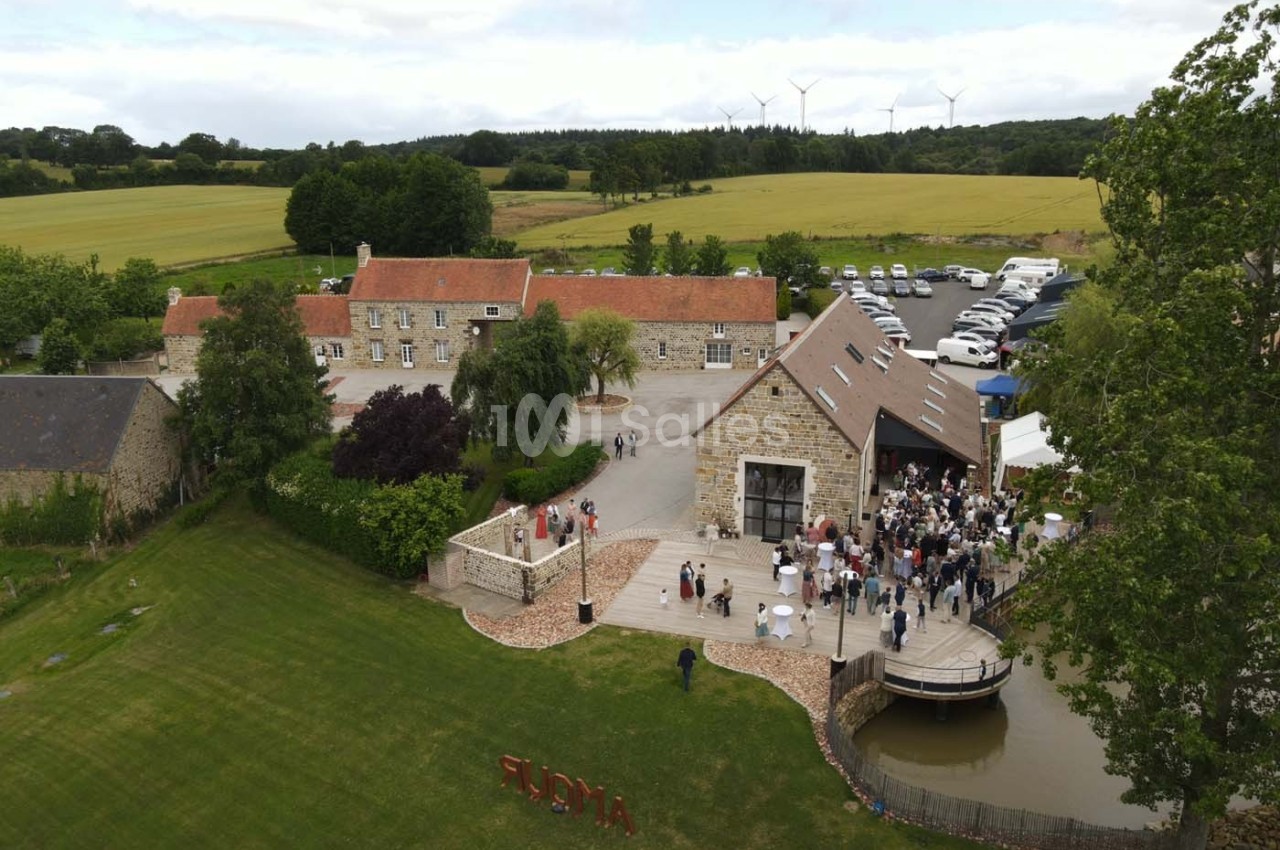 Vue aérienne d'un domaine rural avec bâtiments en pierre, terrasse animée, pelouse et champs environnants.