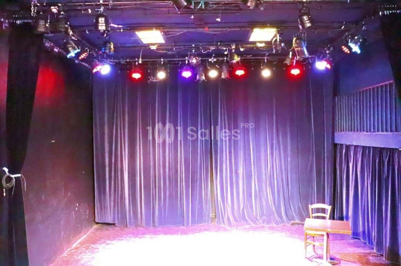 Scène de théâtre vide avec rideaux violets, éclairée par des projecteurs colorés, une chaise et une table en bois.