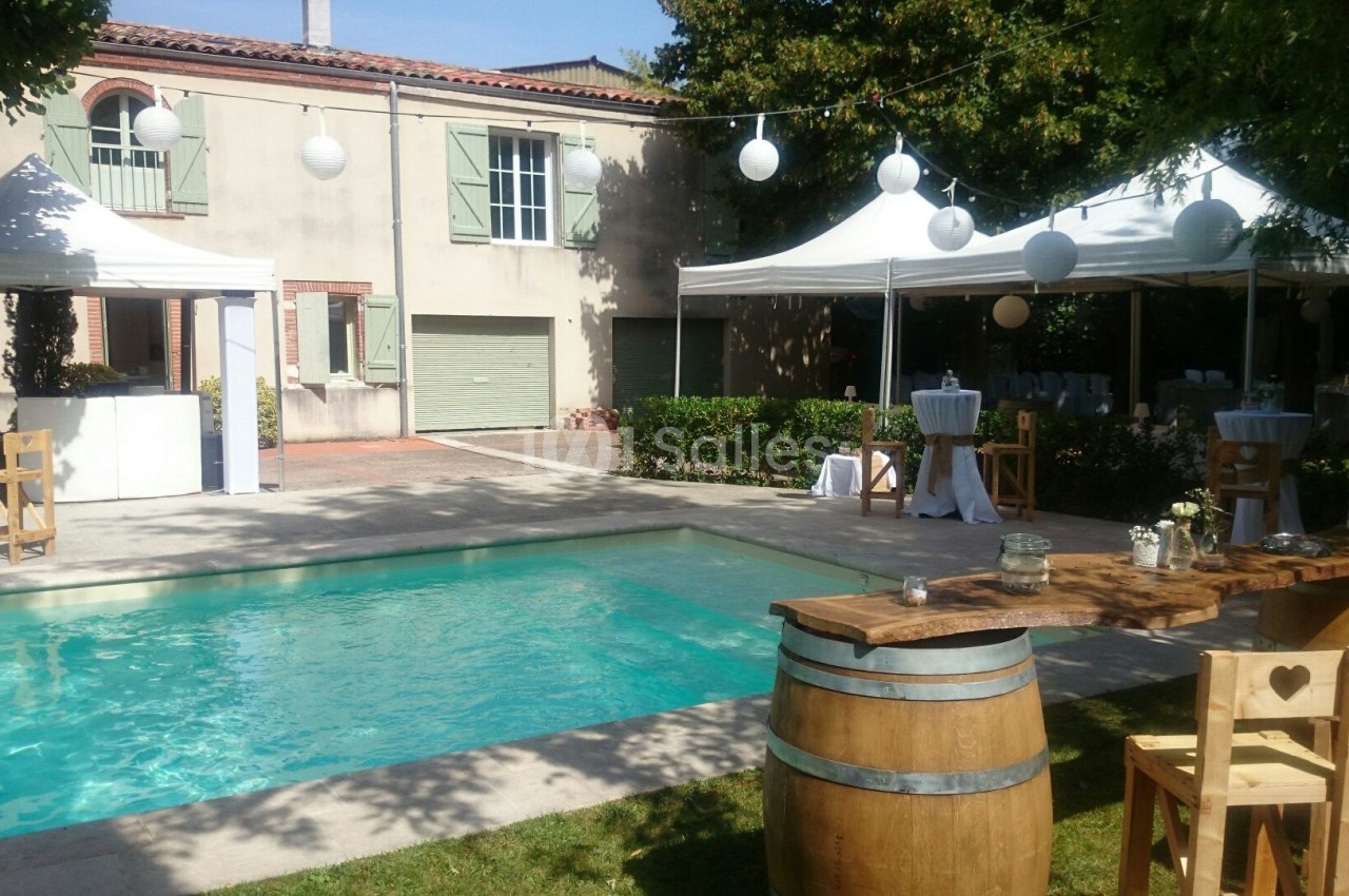 Jardin avec piscine, tables en bois, tonneaux et tentes blanches décorées pour un événement en plein air.