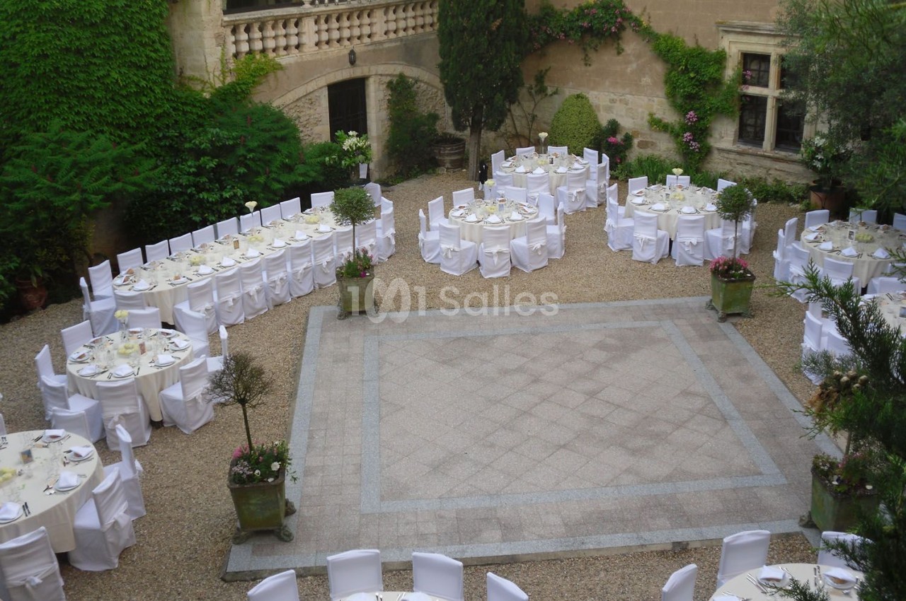 Tables rondes dressées avec nappes blanches dans une cour pavée entourée de murs en pierre et de végétation.