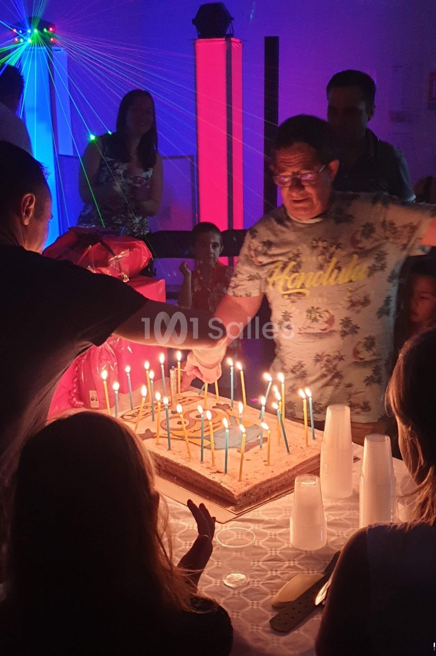 Un homme souffle des bougies sur un grand gâteau d'anniversaire entouré de personnes dans une ambiance festive.