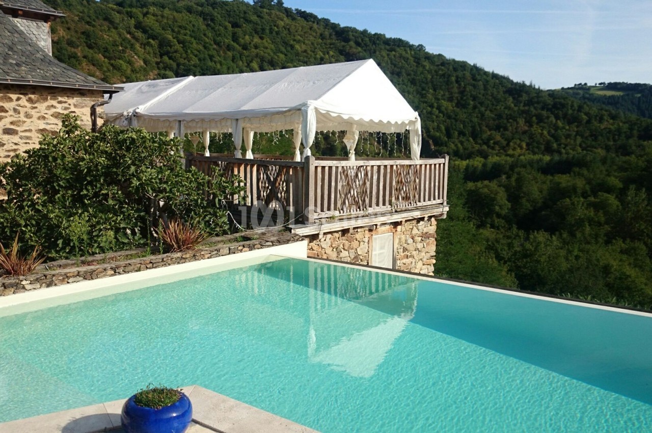 Piscine à débordement avec vue sur une colline boisée, terrasse en bois et tente blanche attenantes.