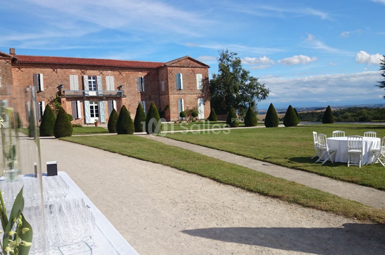 Grande bâtisse en briques rouges entourée de pelouses, avec une table dressée et des chaises blanches en extérieur.