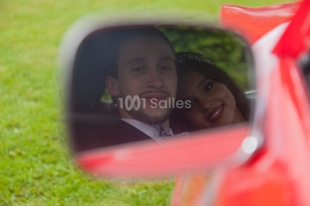 Reflet d'un couple souriant dans le rétroviseur d'une voiture rouge, avec un fond d'herbe verte floue.