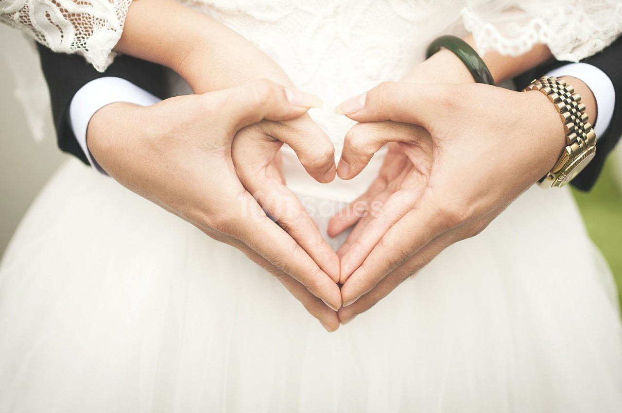 Deux mains formant un cœur devant une robe de mariée, entourées par des mains masculines en costume.