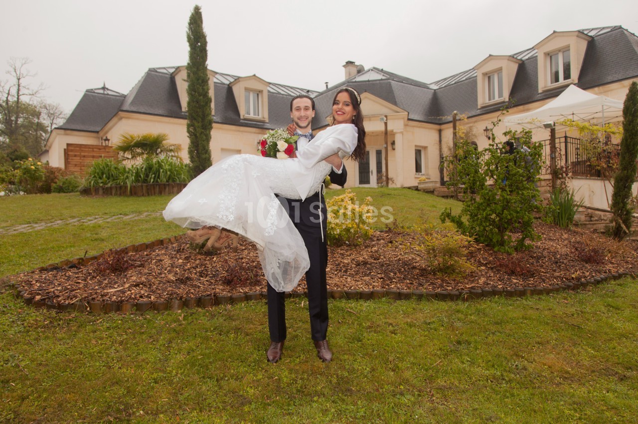 Un homme en costume porte une femme en robe de mariée devant une grande maison avec jardin.