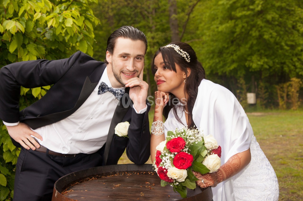 Un couple en tenue de mariage pose près d'un tonneau en bois dans un jardin verdoyant.