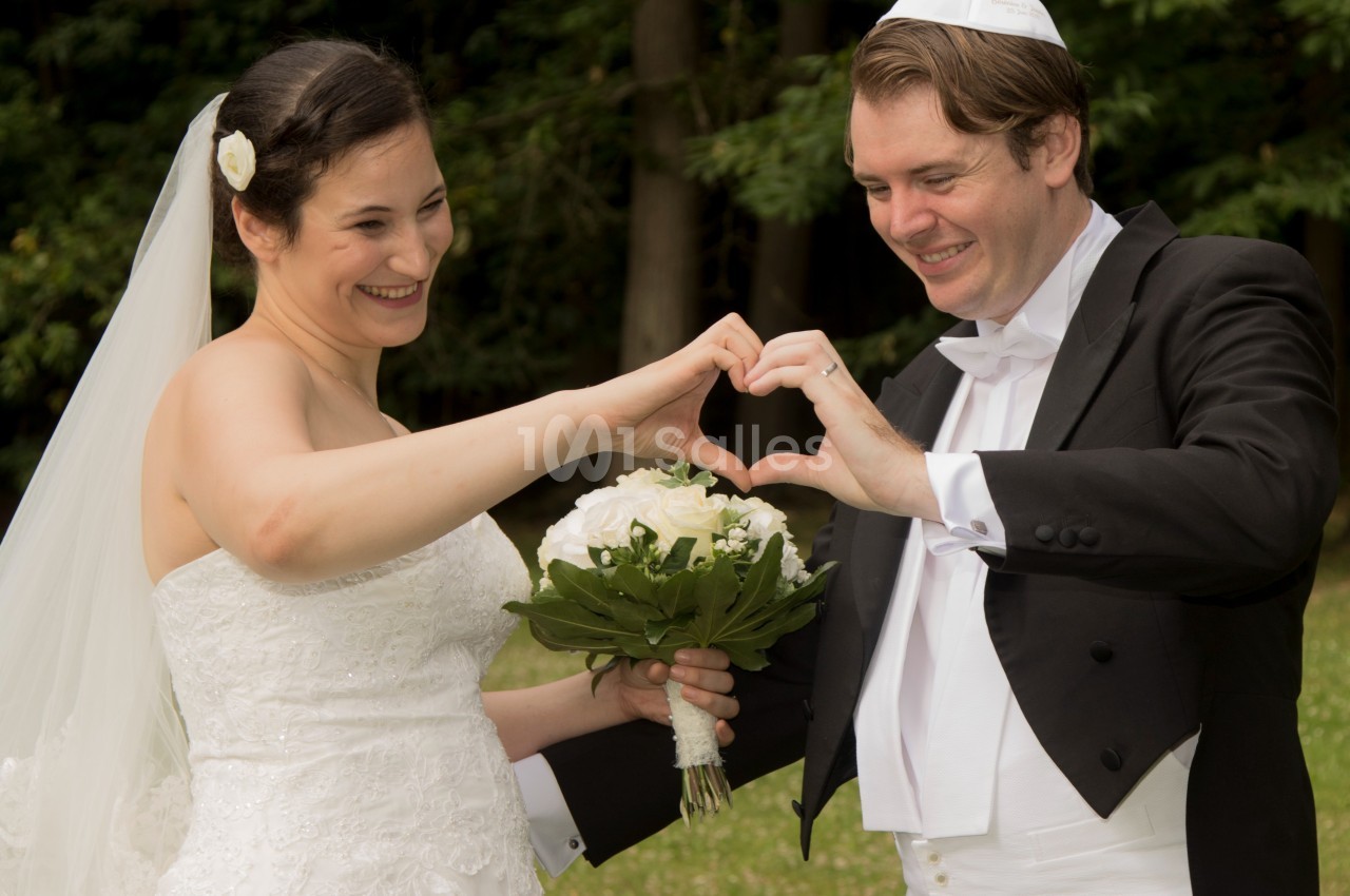 Un couple de mariés souriants formant un cœur avec leurs mains, tenant un bouquet de fleurs blanches, en extérieur.