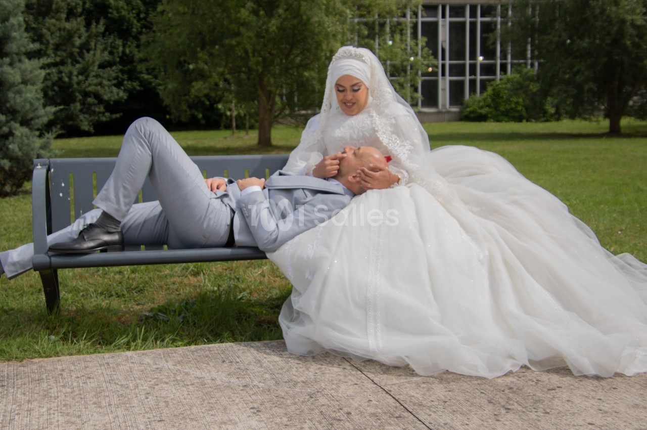 Un couple en tenue de mariage, la mariée assise sur un banc tenant la tête du marié allongé sur ses genoux, dans un parc.
