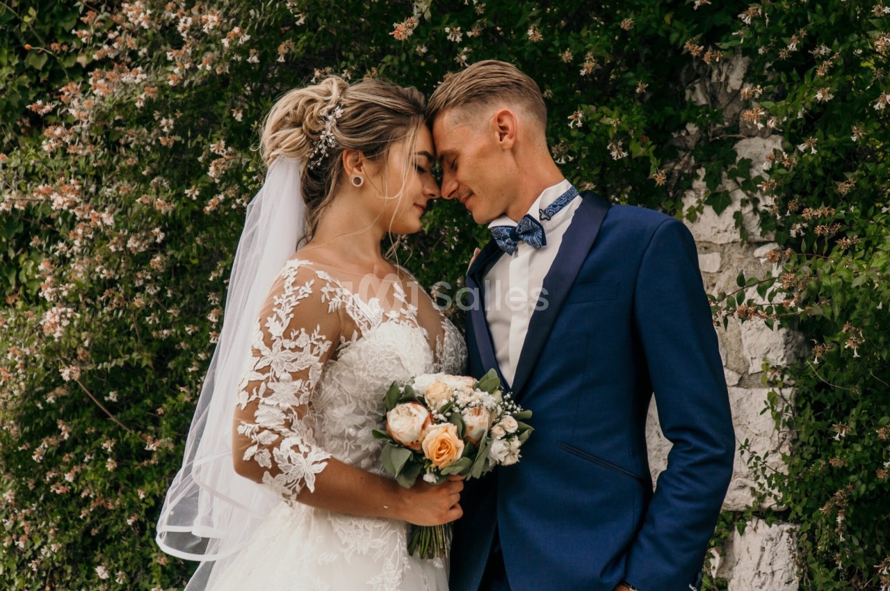 Un couple de mariés en tenue de cérémonie se tient face à face devant un mur fleuri.