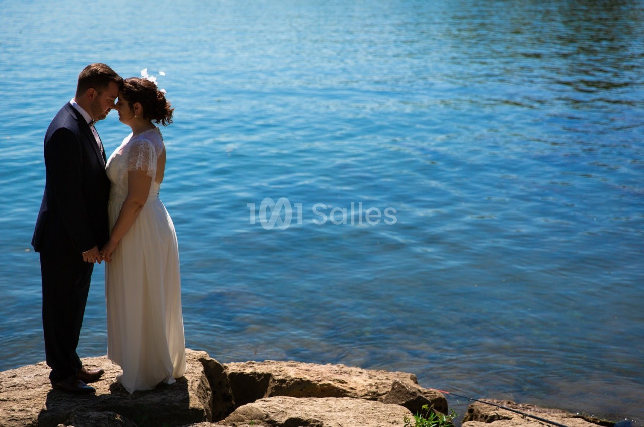 Un couple en tenue de mariage se tient la main sur des rochers au bord d'un lac sous un ciel dégagé.