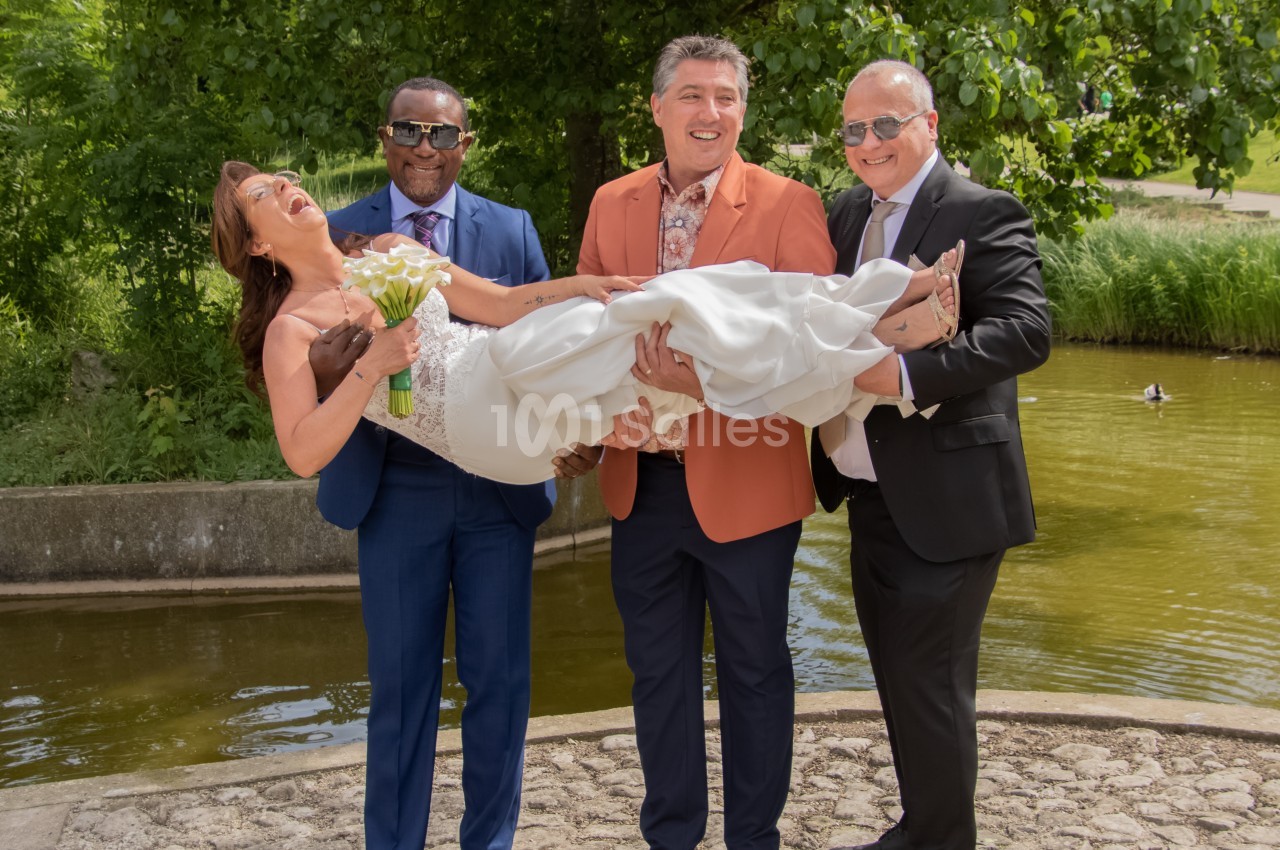 Une mariée souriante en robe blanche est portée par trois hommes élégants devant un étang et des arbres.