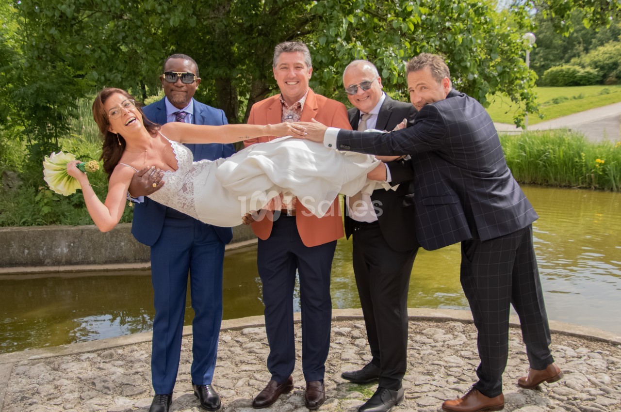 Une mariée en robe blanche est portée en l'air par quatre hommes souriants dans un parc verdoyant près d'un étang.