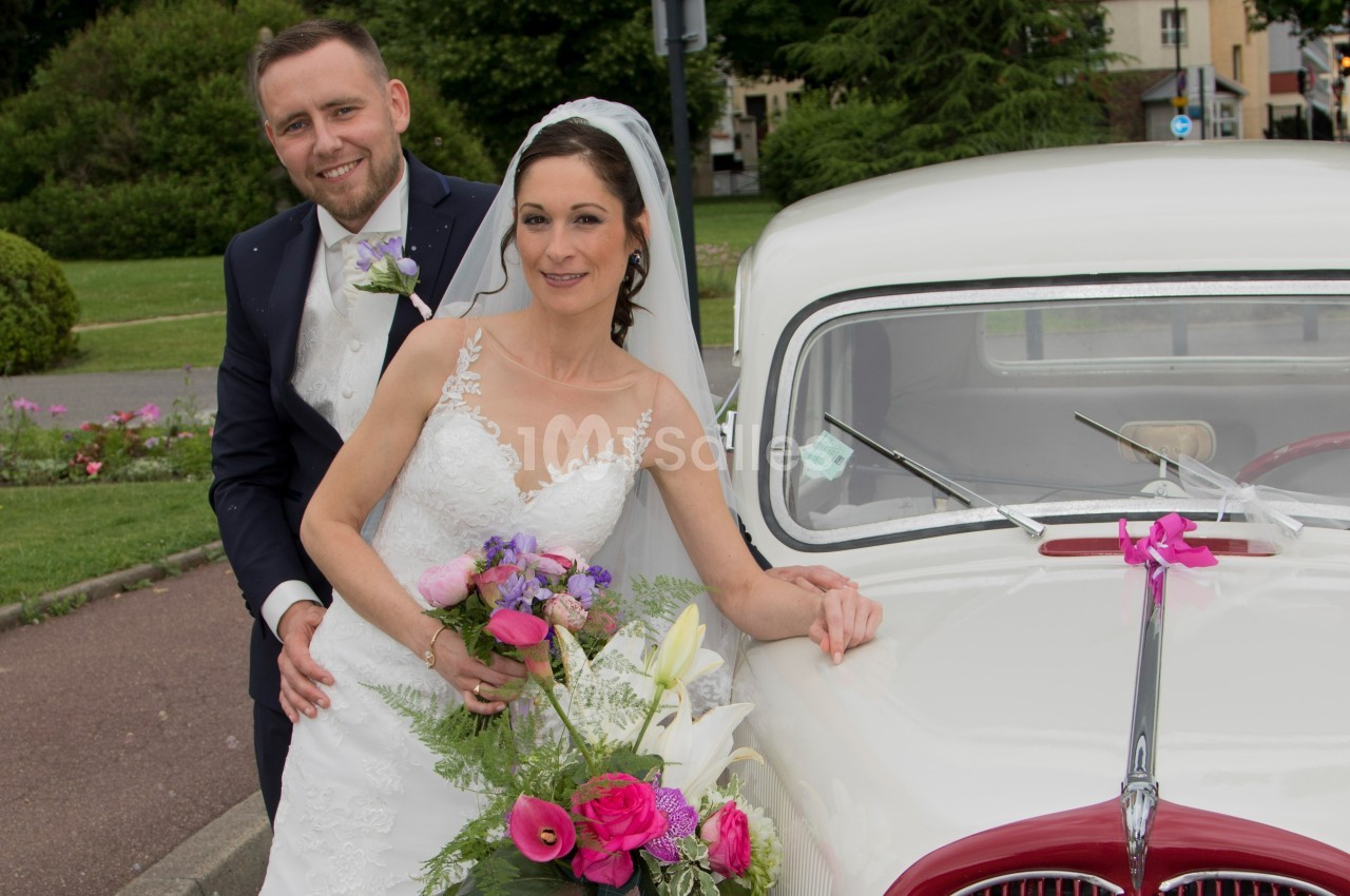 Un couple de mariés pose souriant près d'une voiture ancienne blanche, avec un bouquet de fleurs colorées.