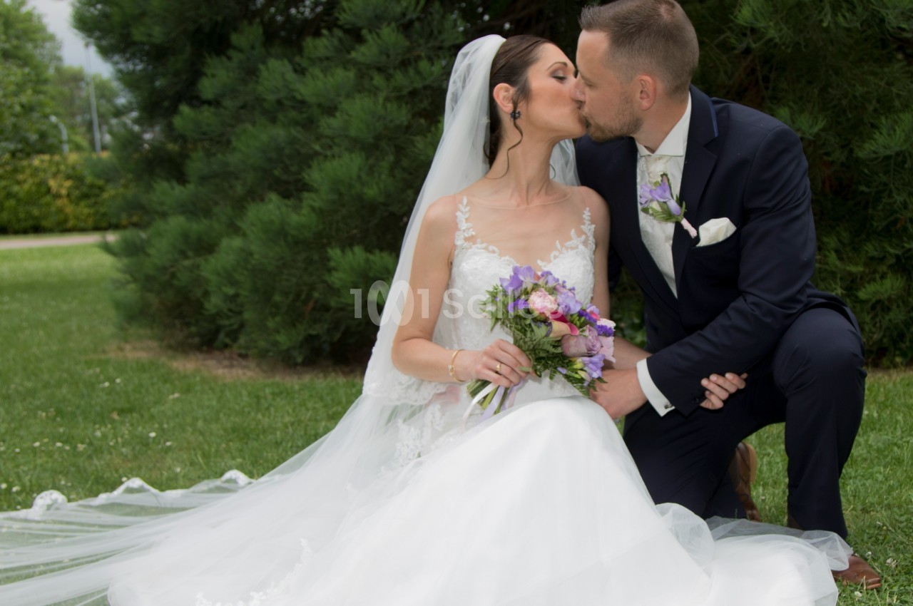Un couple de mariés s'embrasse dans un jardin, la mariée en robe blanche tenant un bouquet de fleurs colorées.