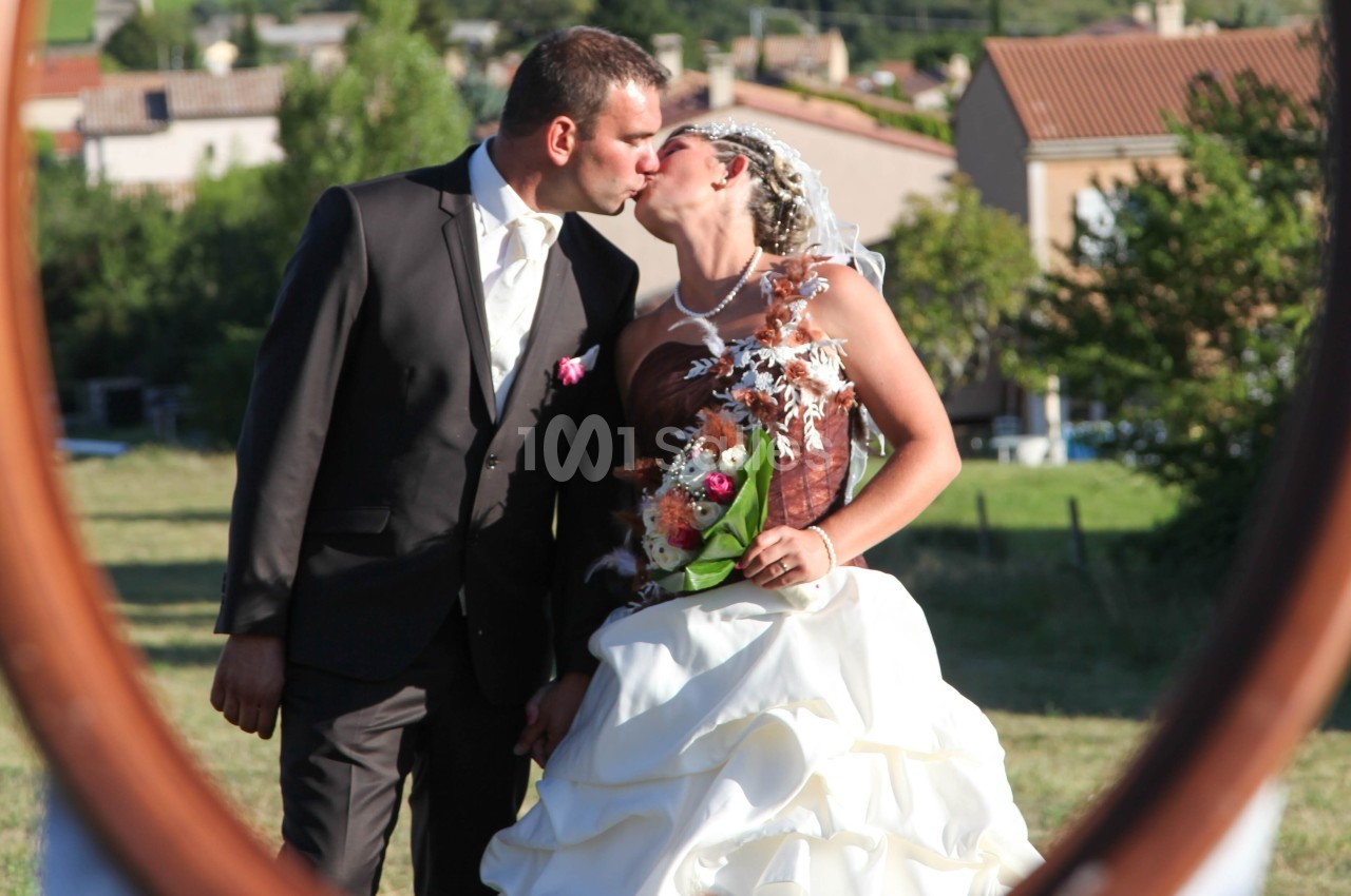 Un couple en tenue de mariage s'embrasse dans un cadre extérieur ensoleillé avec des maisons en arrière-plan.
