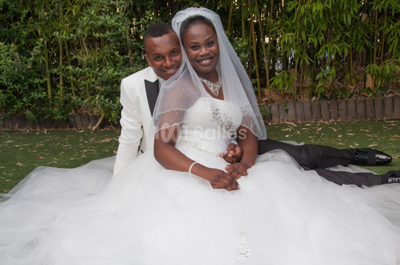 Un couple de mariés souriant, assis sur une pelouse, la mariée en robe blanche et le marié en costume clair.