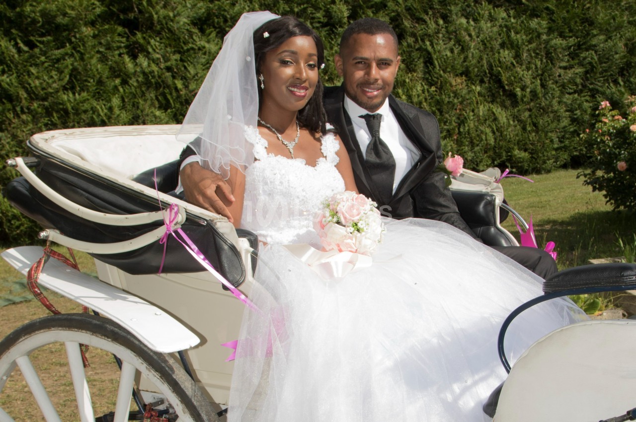 Un couple en tenue de mariage assis dans une calèche blanche décorée de rubans roses, en extérieur.