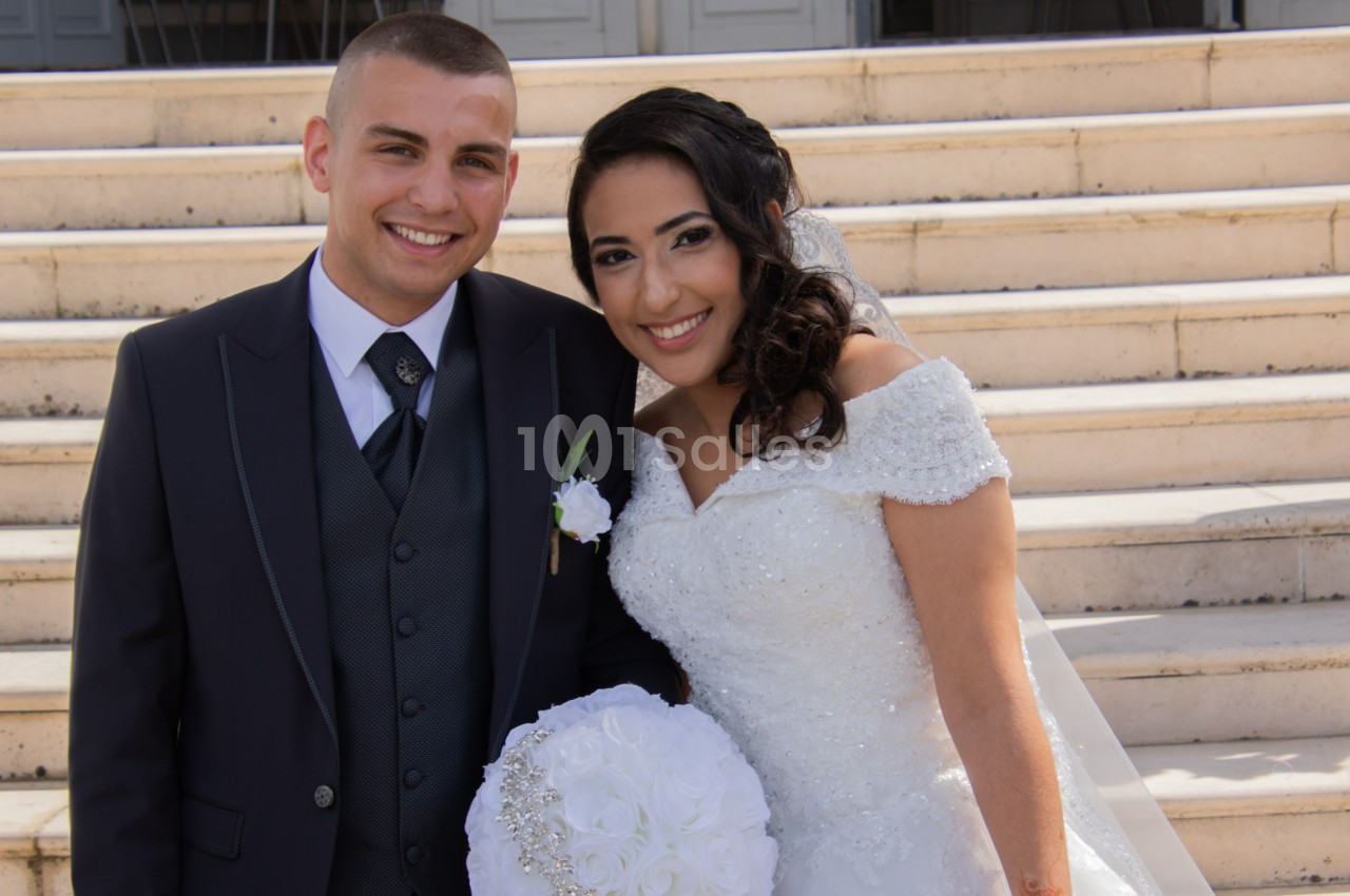 Un couple souriant en tenue de mariage pose devant des marches en pierre.