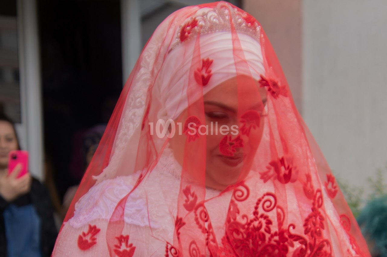 Femme portant un voile rouge orné de motifs floraux, vêtue de blanc, lors d'une cérémonie ou d'un événement festif.