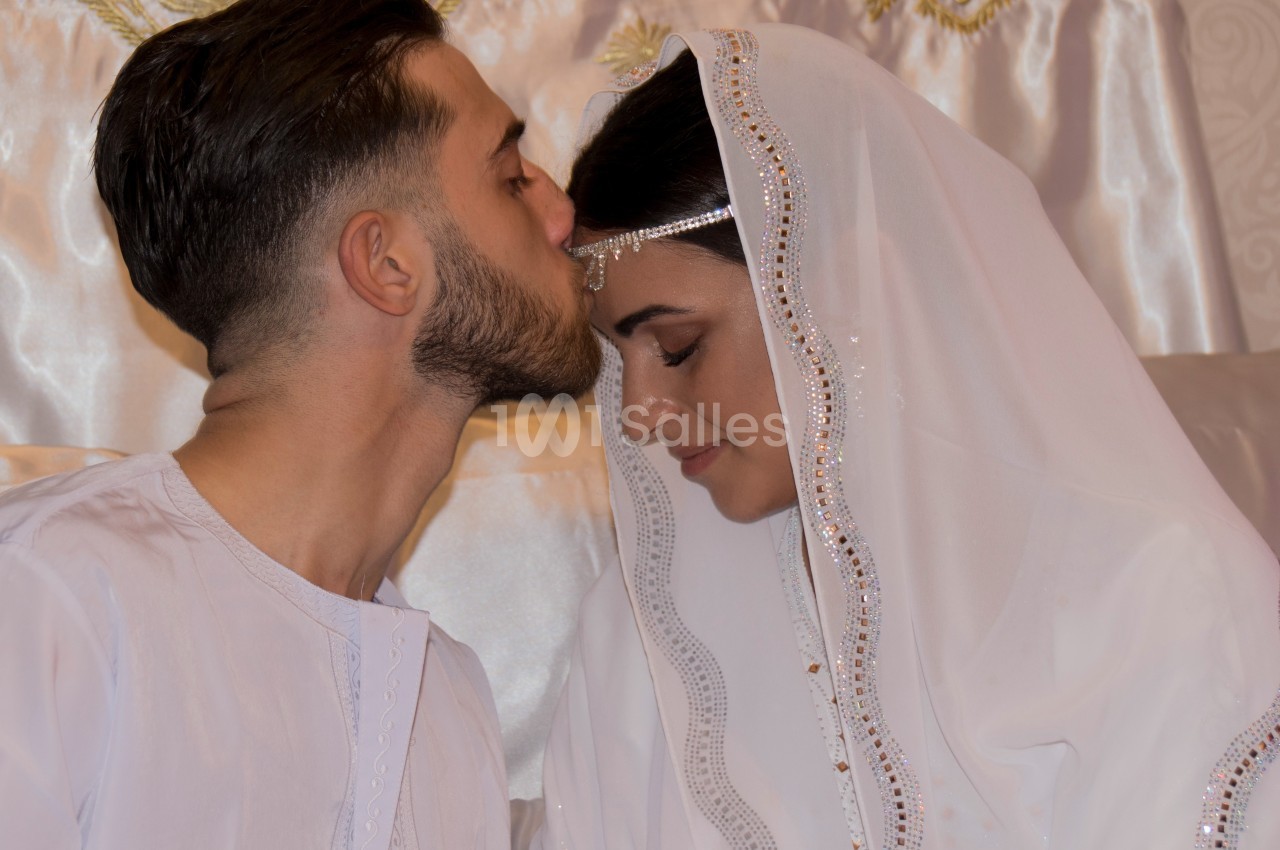 Un homme embrasse tendrement le front d'une femme portant un voile blanc orné, dans un cadre intime et lumineux.