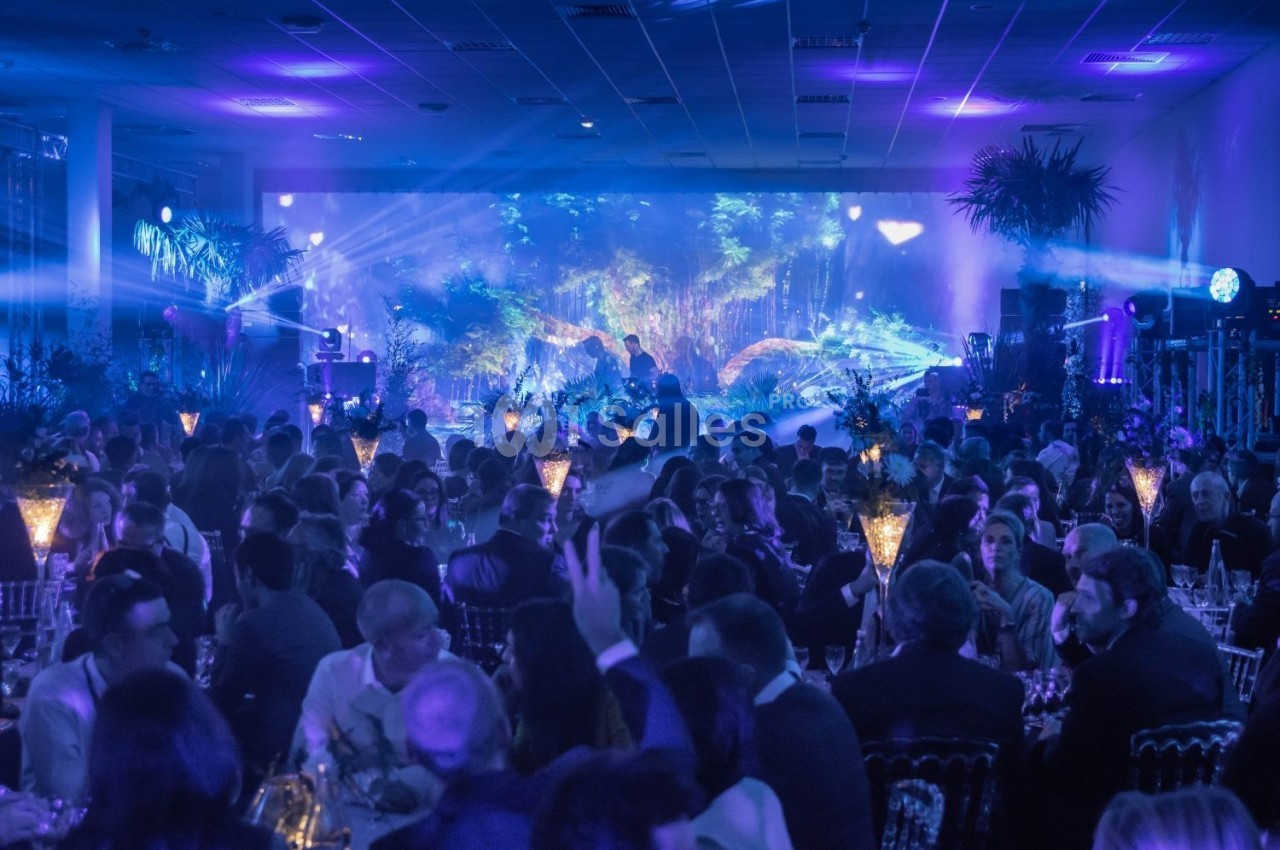 Salle de réception illuminée en bleu avec des invités assis à des tables et une scène décorée en arrière-plan.