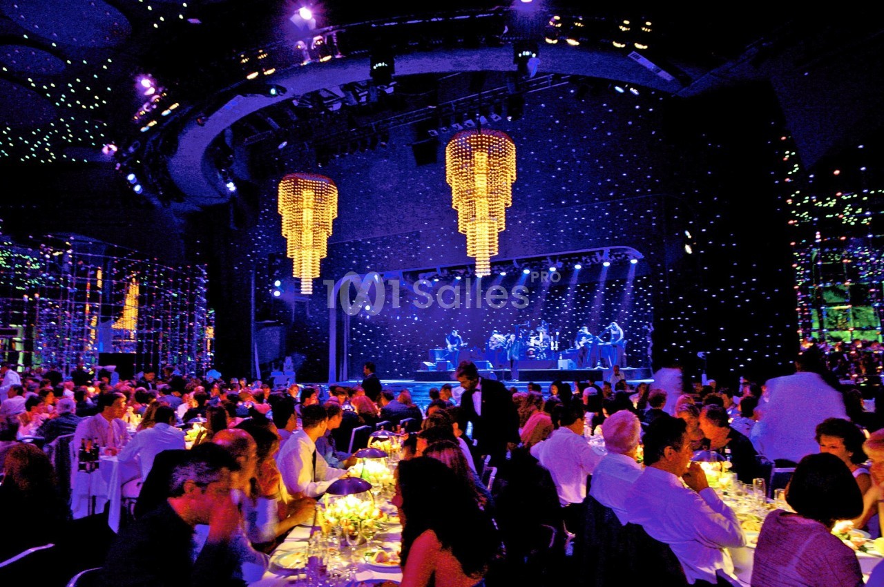 Salle de spectacle illuminée avec des convives dînant devant une scène où des musiciens jouent sous des lustres suspendus.
