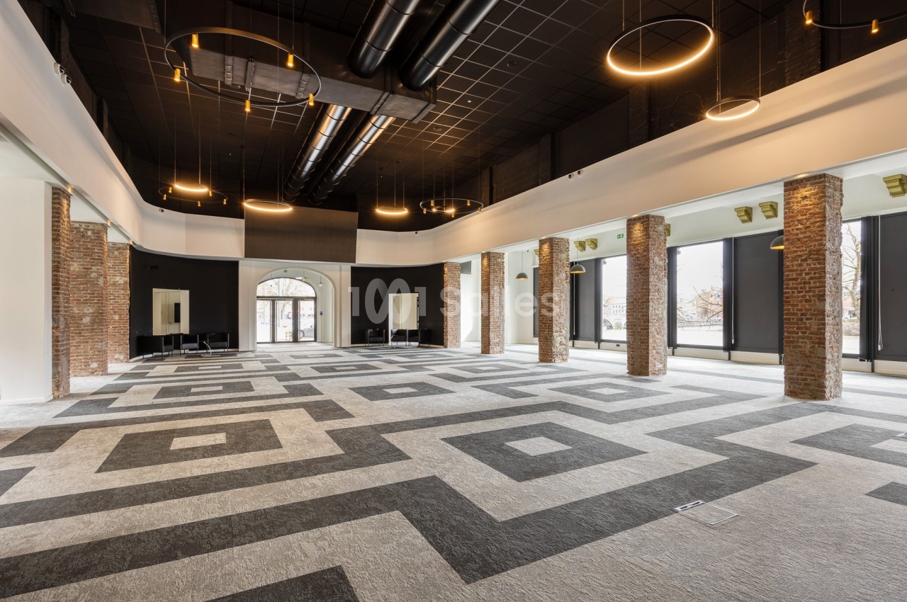 Salle spacieuse avec sol géométrique, colonnes en briques, grandes fenêtres et plafonniers circulaires modernes.
