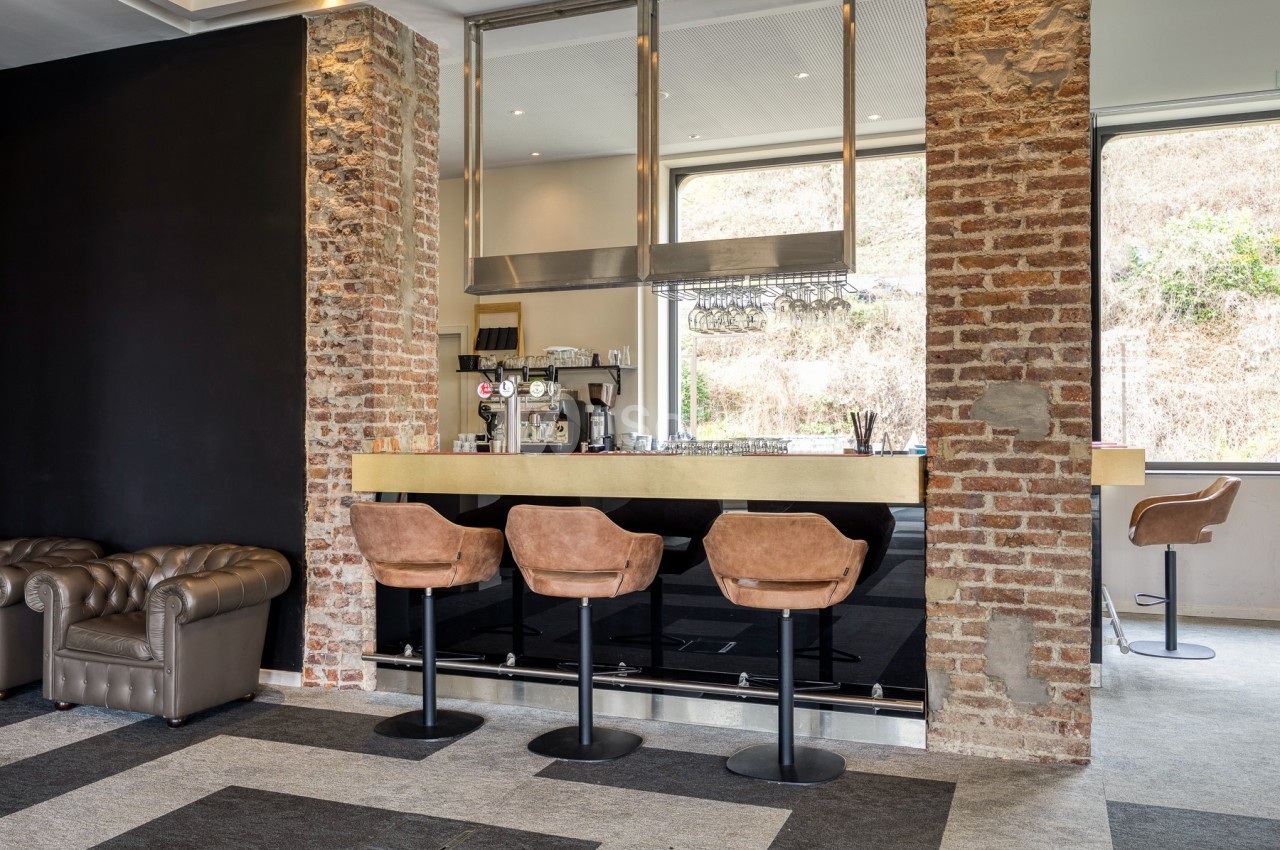 Bar moderne avec comptoir en bois, tabourets en cuir, murs en briques apparentes et grandes fenêtres lumineuses.