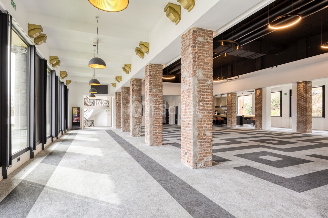 Salle lumineuse avec colonnes en briques, grandes fenêtres, sol en motifs géométriques et luminaires suspendus.