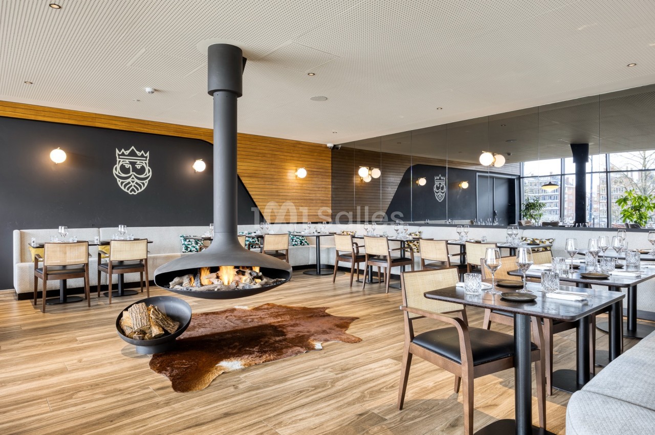 Salle de restaurant moderne avec cheminée suspendue, tables dressées et décoration épurée sur fond de murs sombres.