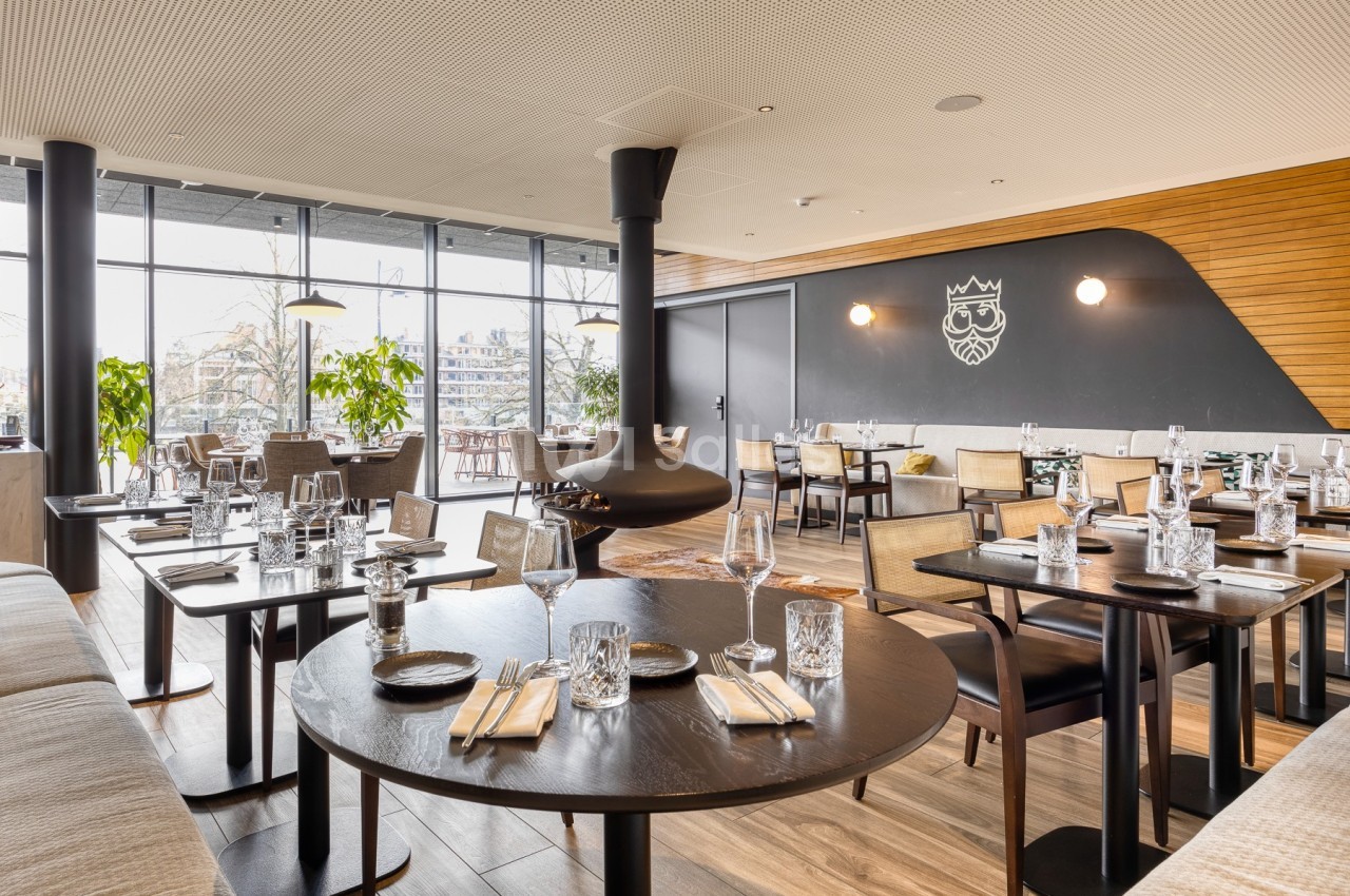 Salle de restaurant moderne avec tables dressées, grandes baies vitrées et cheminée suspendue au centre.