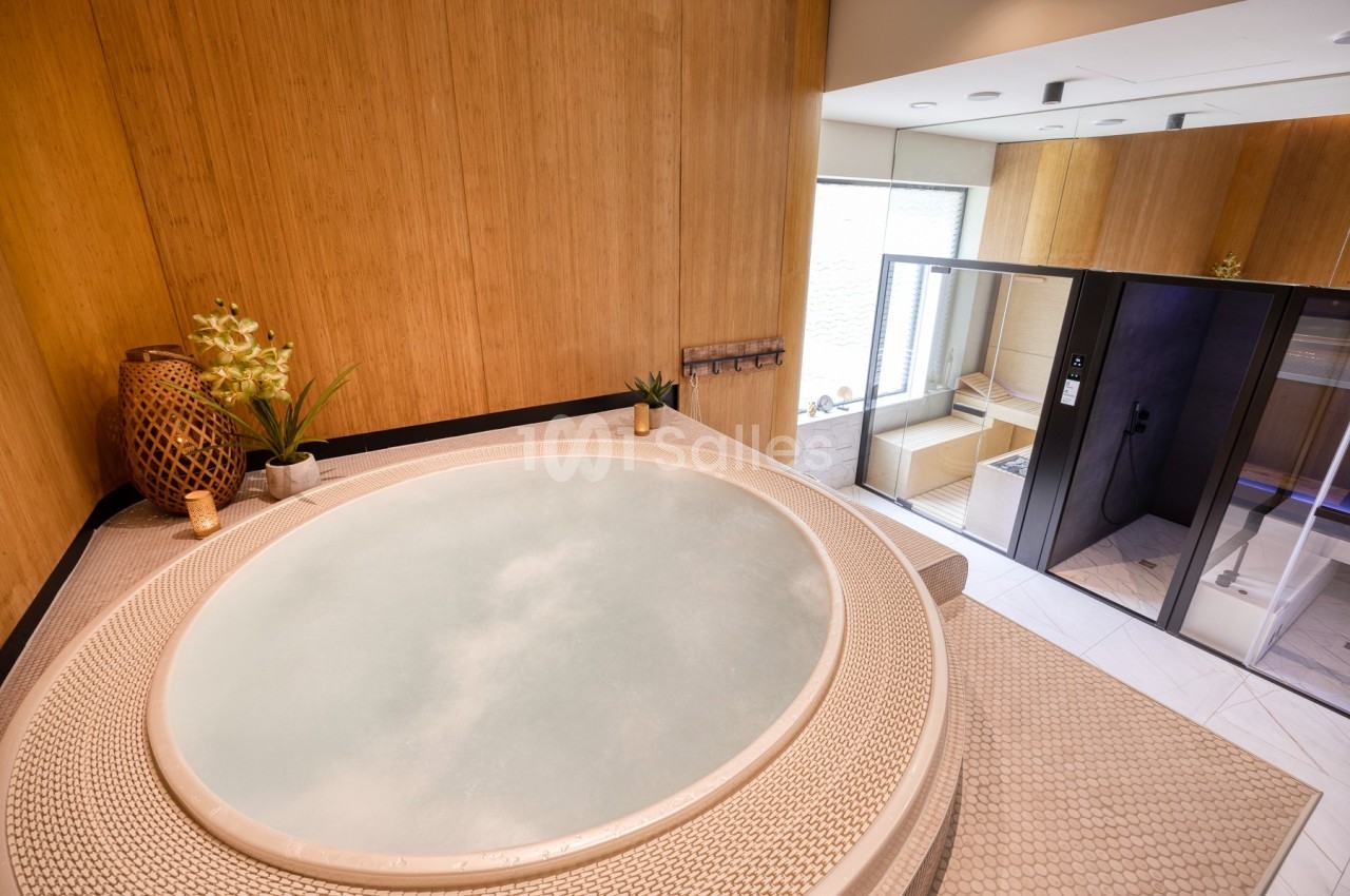 Bain à remous circulaire dans une pièce en bois clair avec une douche et un sauna vitré en arrière-plan.