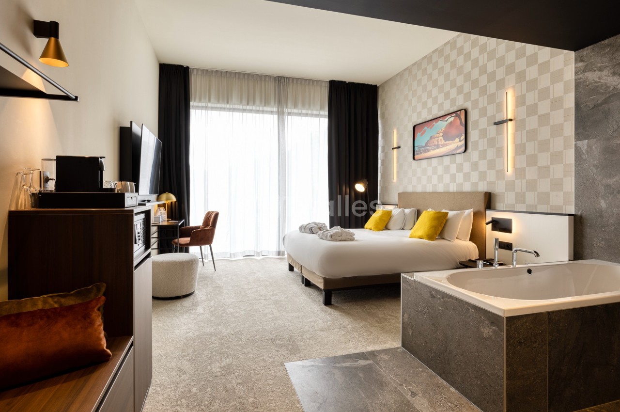 Chambre d'hôtel moderne avec lit double, baignoire intégrée, bureau, télévision et grande baie vitrée avec rideaux.