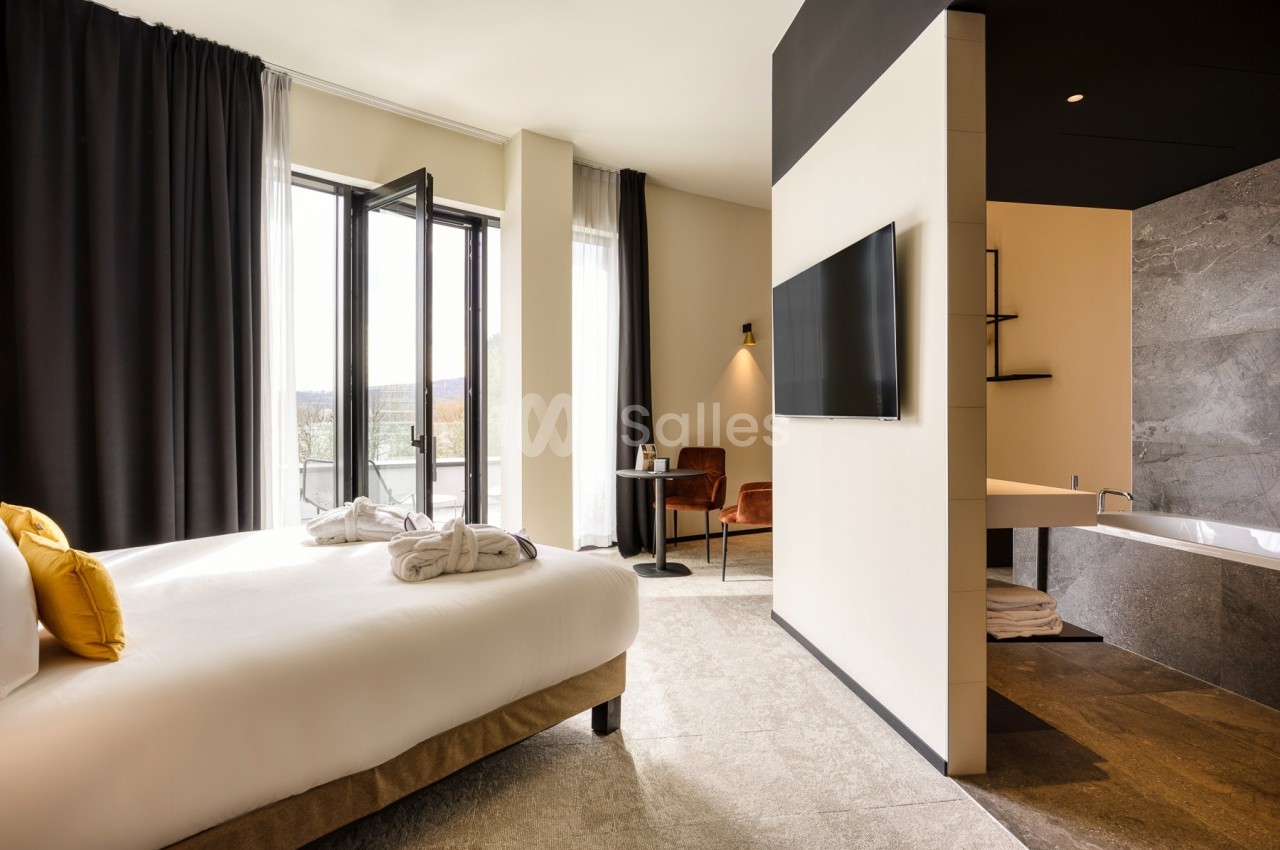 Chambre lumineuse avec lit double, télévision murale, salle de bain ouverte et balcon avec vue extérieure.