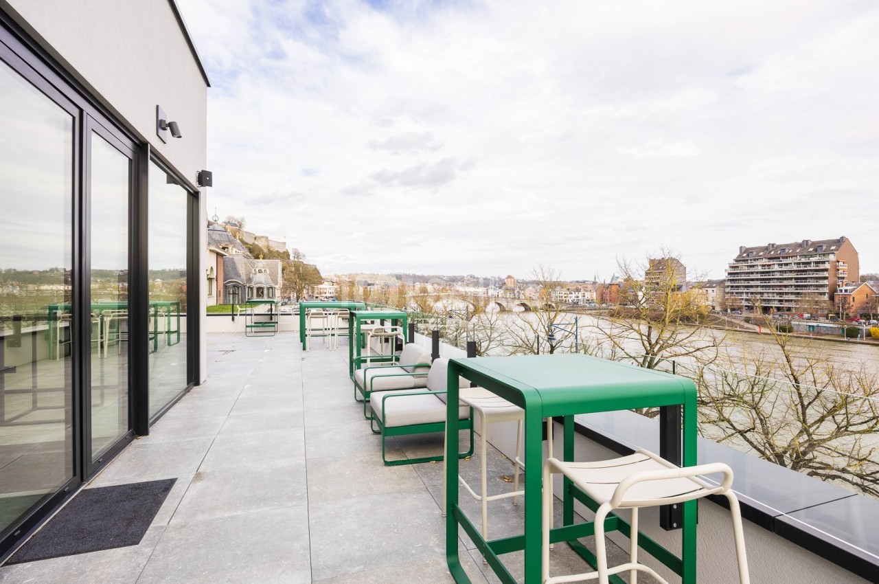 Terrasse avec mobilier moderne, vue sur une rivière bordée d'arbres et de bâtiments résidentiels.