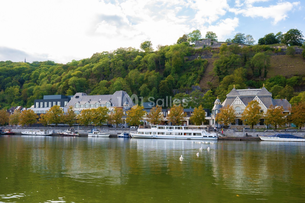 Vue sur une rivière bordée de bateaux, avec des bâtiments historiques et une colline boisée en arrière-plan.