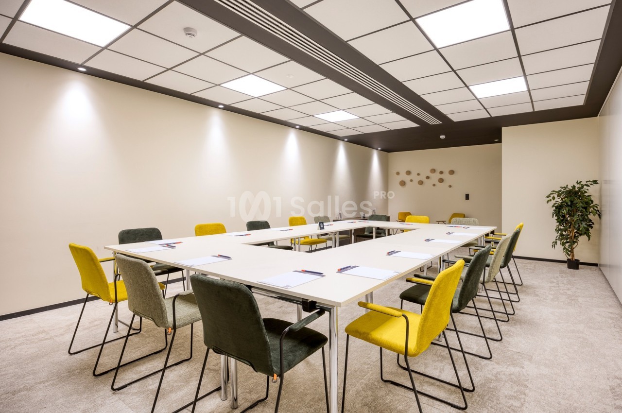 Salle de réunion lumineuse avec tables disposées en U, chaises colorées et décoration murale minimaliste.