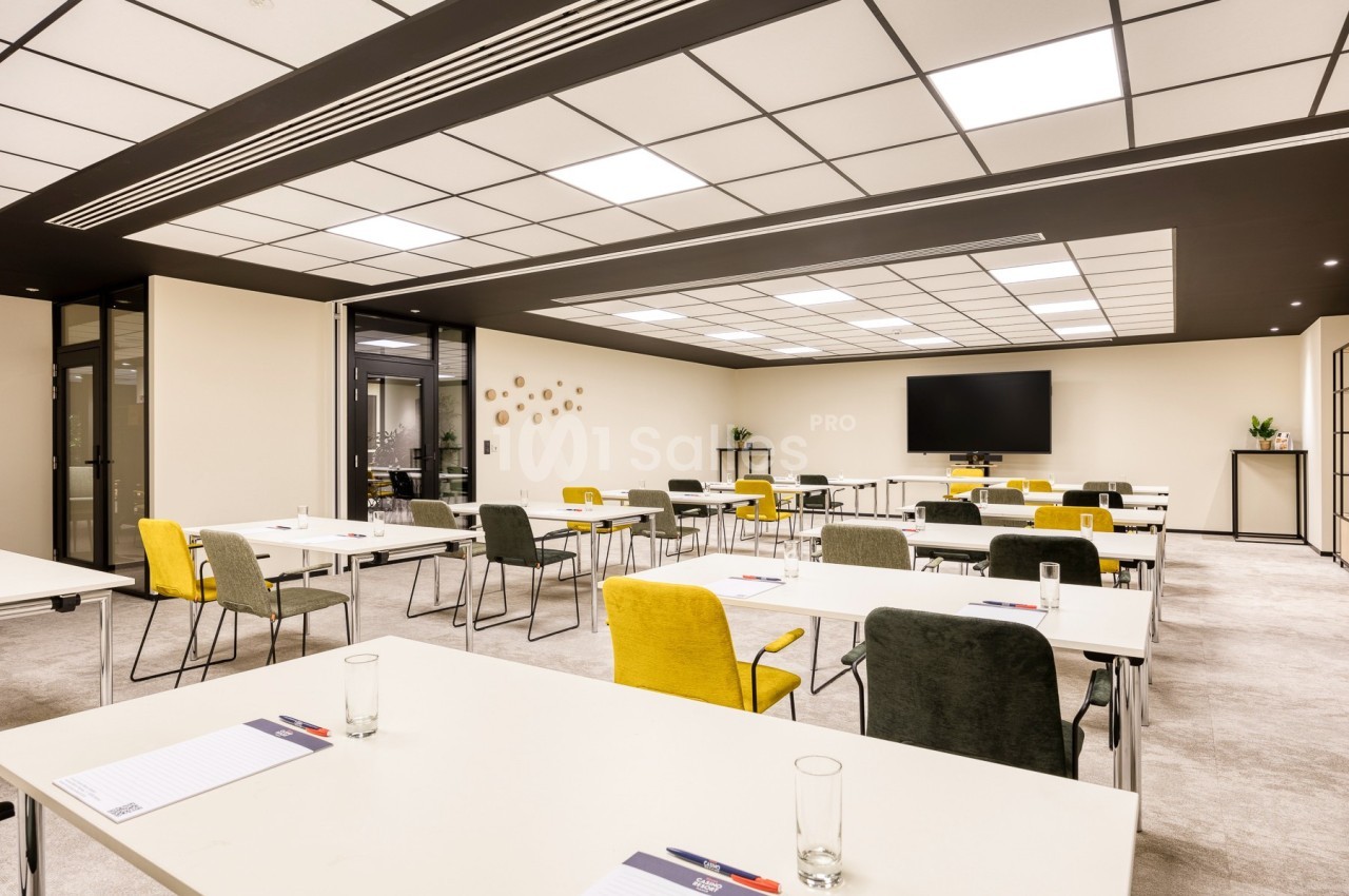 Salle de réunion lumineuse avec tables blanches, chaises colorées, écran mural et décoration minimaliste.