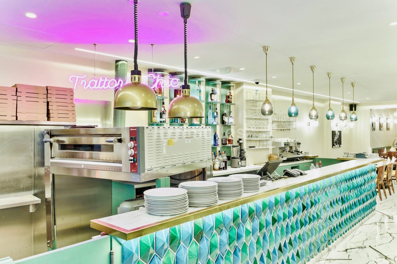 Comptoir de restaurant avec carrelage coloré, four à pizza, étagères garnies et éclairage suspendu.