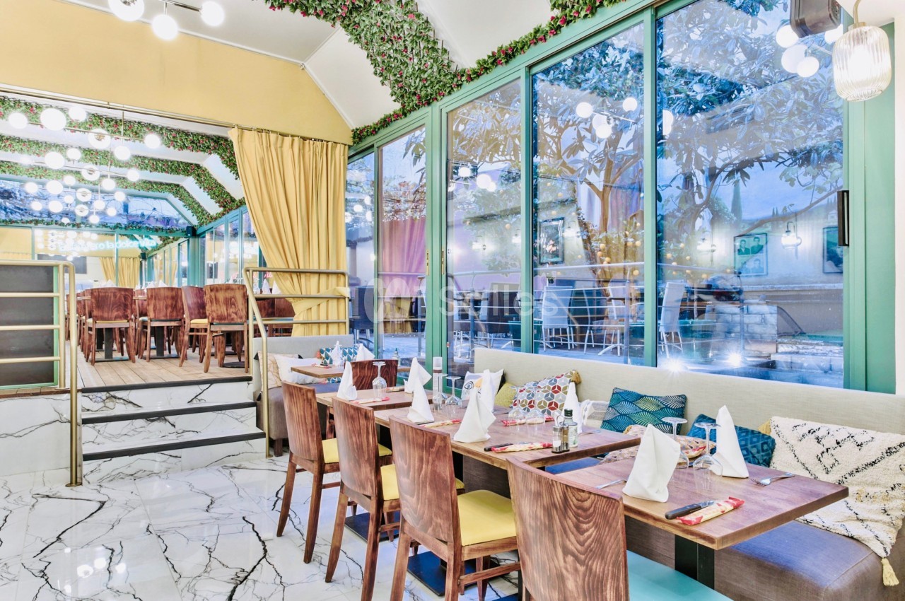 Salle de restaurant lumineuse avec tables dressées, grandes baies vitrées et décoration végétale au plafond.