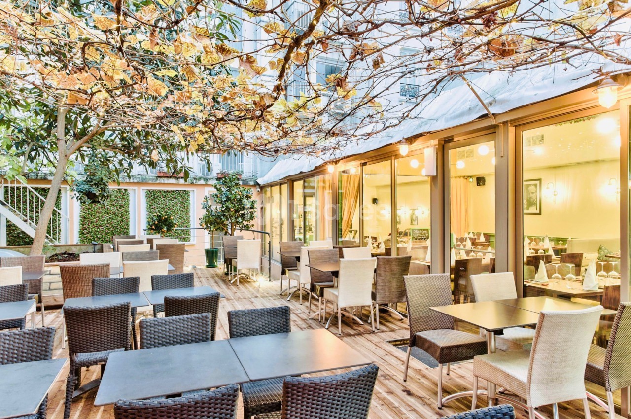 Terrasse d'un restaurant avec tables et chaises en osier, entourée de végétation et éclairée par des guirlandes lumineuses.