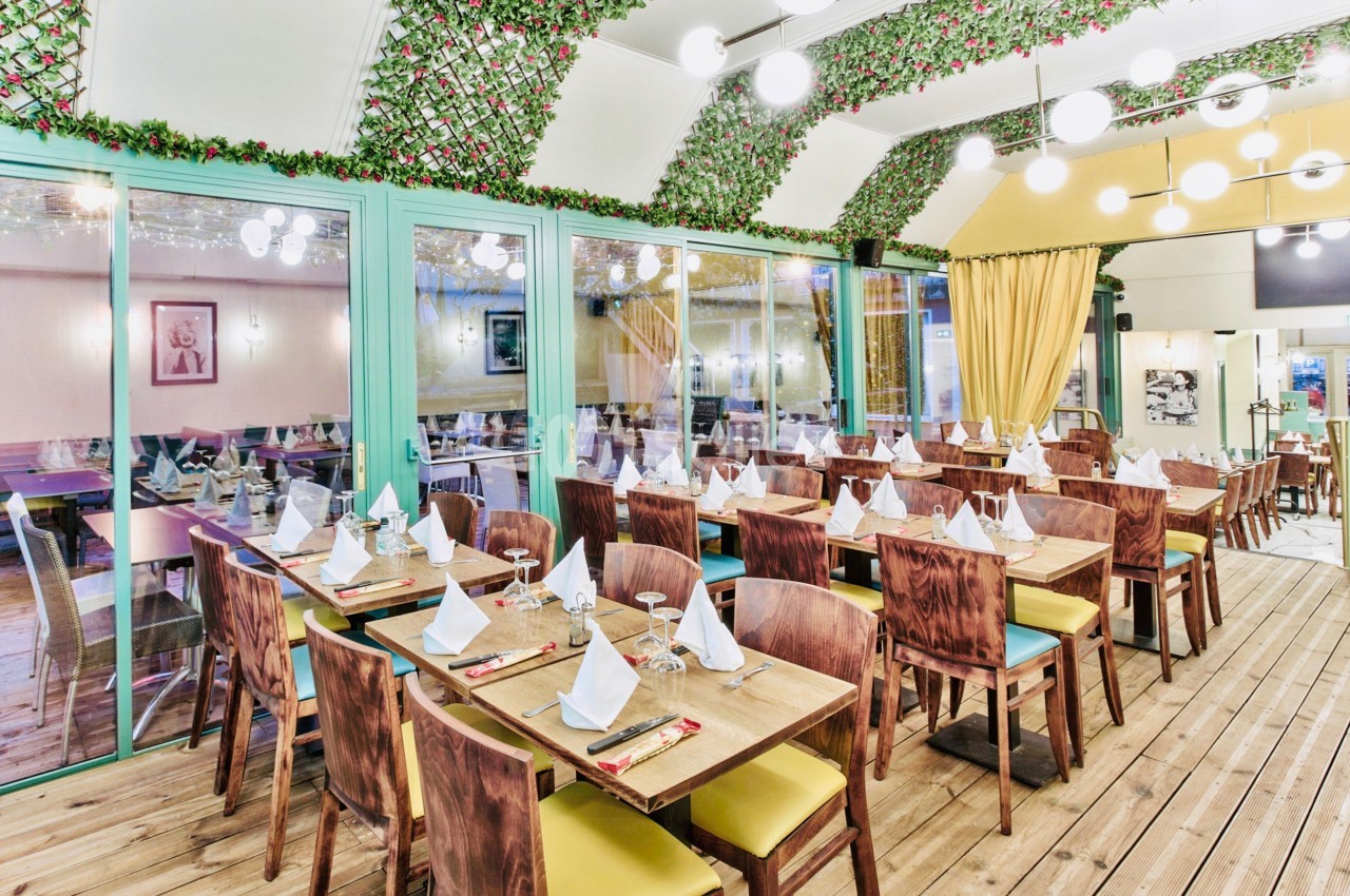 Salle de restaurant lumineuse avec tables en bois dressées, décorée de guirlandes végétales et éclairée par des suspensions.