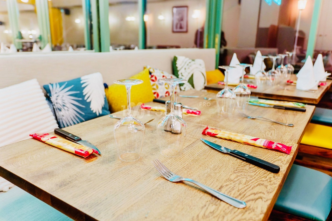 Table dressée avec des couverts, verres à pied et serviettes, entourée de coussins colorés dans un restaurant lumineux.