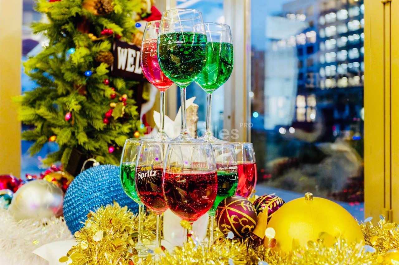 Verres colorés empilés en pyramide devant une fenêtre, décorations de Noël et guirlandes en arrière-plan.